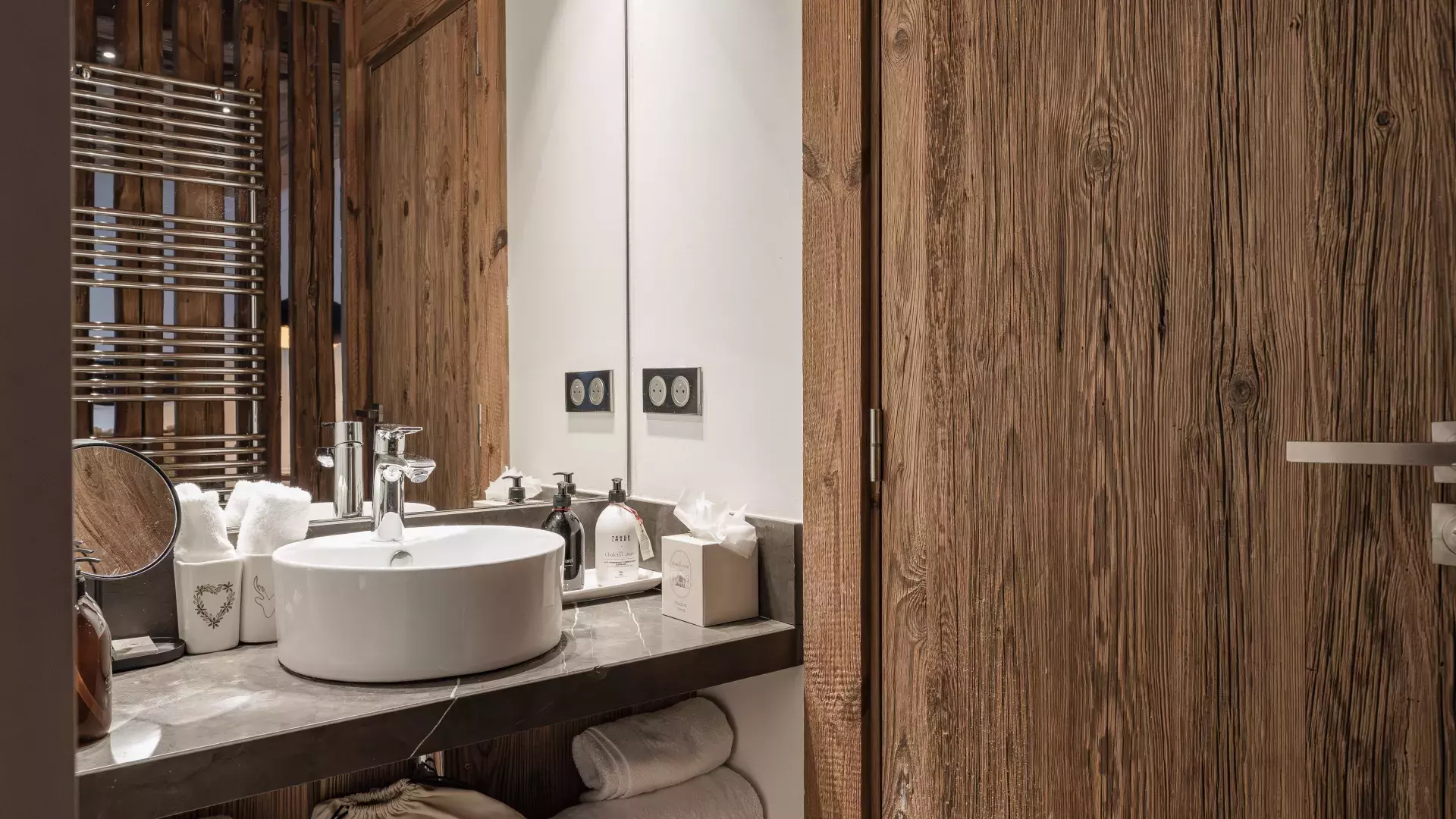 Détail d'une salle de bain avec vasque ronde sur plan en pierre, grand miroir et finitions en vieux bois au Penthouse Savoie.