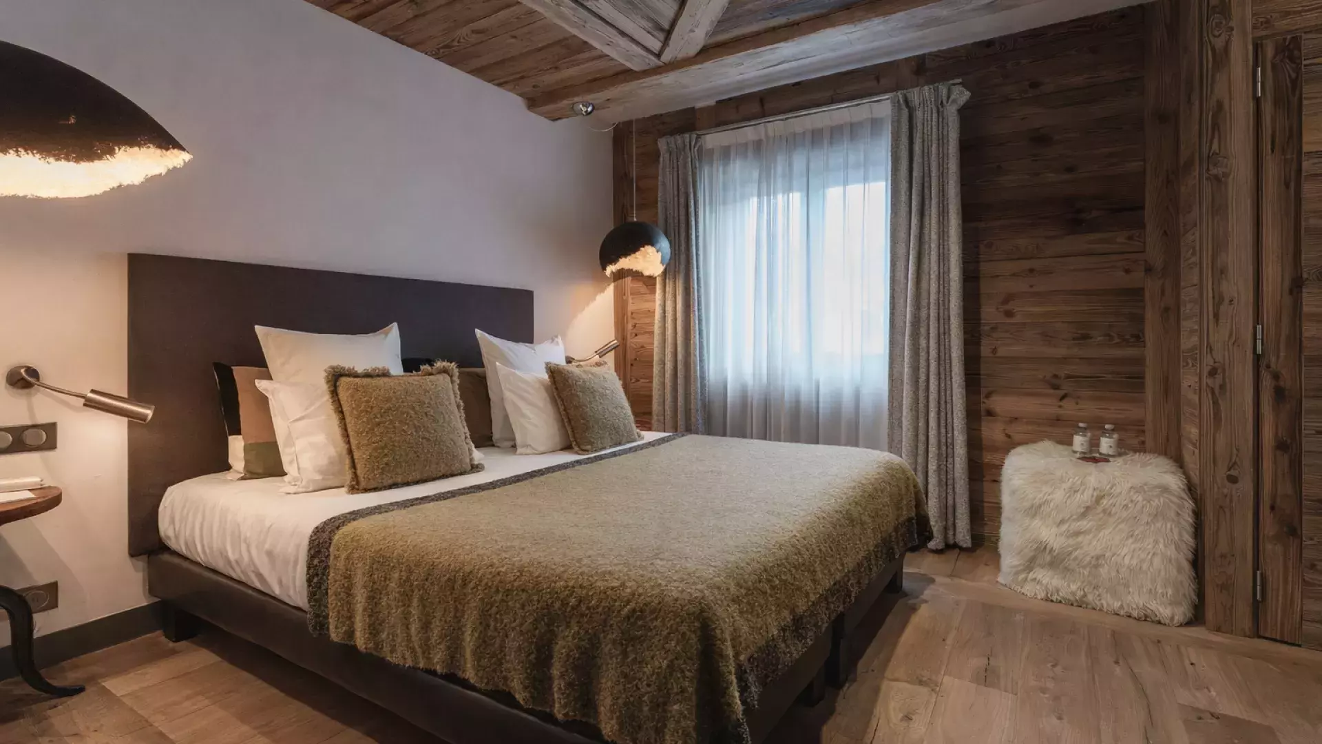 Chambre double élégante aux tons kaki et bois, ambiance cocooning avec suspensions modernes et pouf en fourrure au Penthouse Savoie.