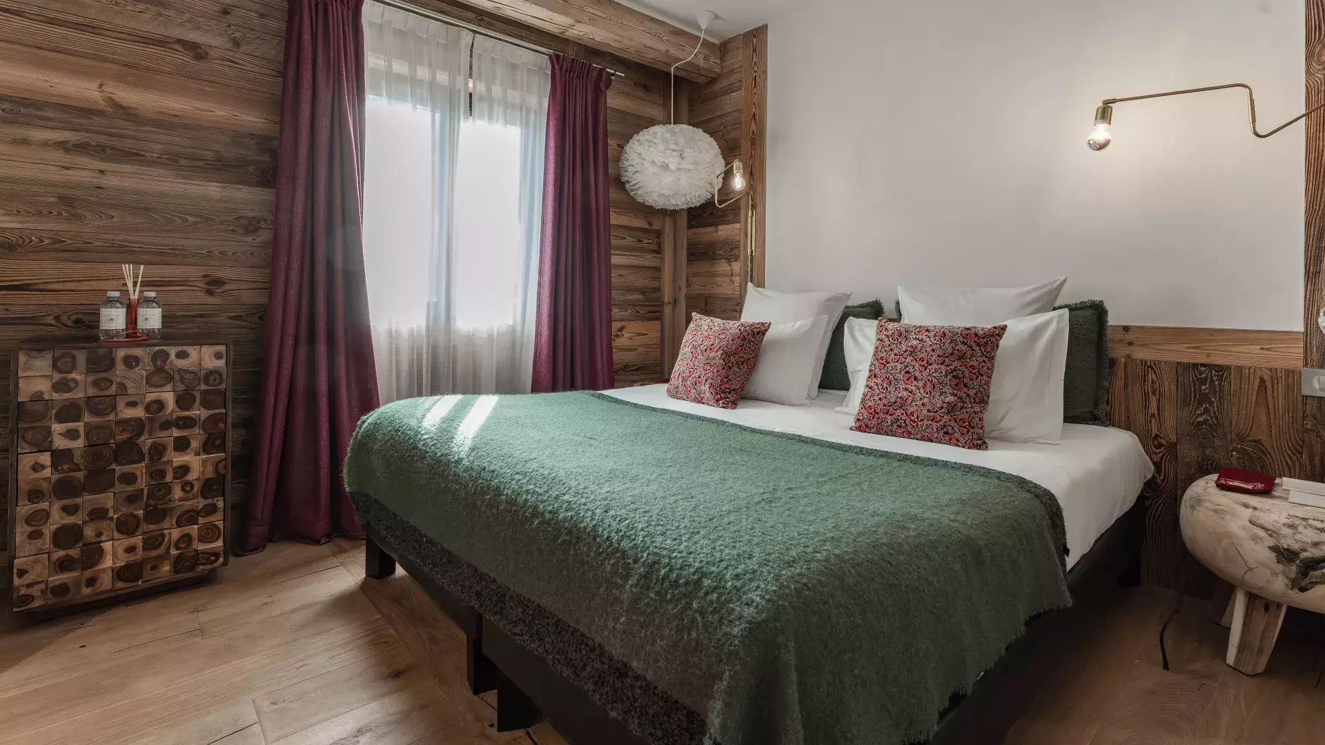 Chambre double chaleureuse aux tons vert et bordeaux, avec commode en bois atypique et suspension plume au Penthouse Savoie.