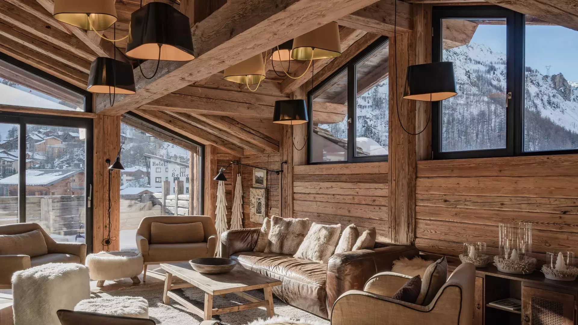 Salon cathédrale sous charpente apparente avec canapé en cuir, multiples suspensions design et vue panoramique sur les sommets de Val d'Isère