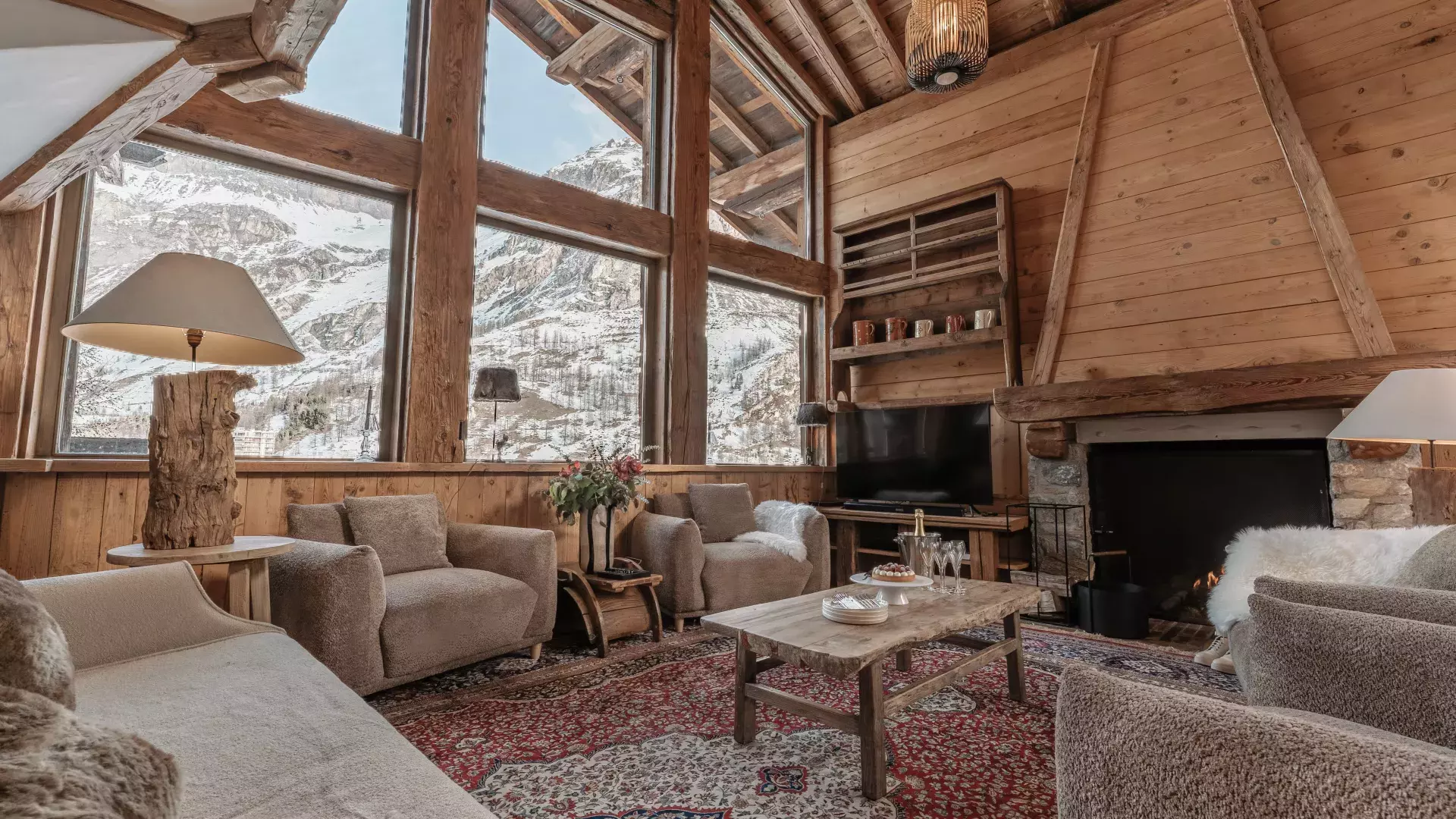 Grand salon lumineux avec immenses baies vitrées face à la montagne, cheminée en pierre massive et tapis d'orient au Chalet Alice.
